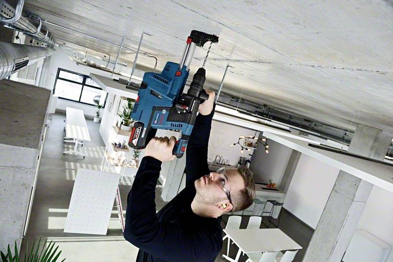 A man wearing safety glasses and ear protection is drilling into the ceiling of a modern office with a blue drill.