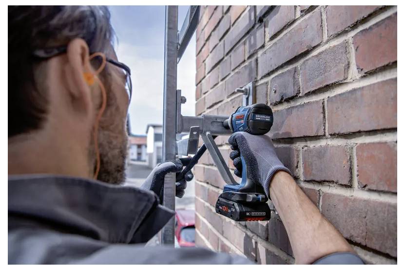 A person wearing gloves and ear protection uses a cordless drill on a brick wall, focusing intently on the task.