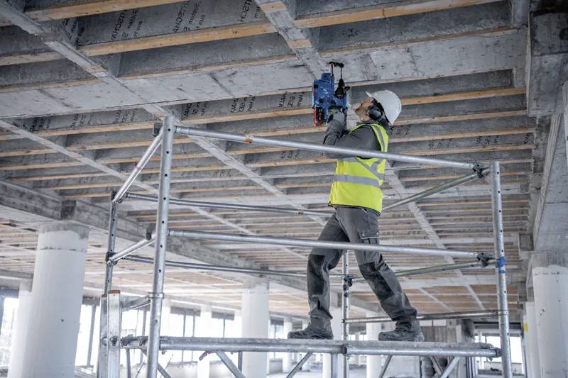 A construction worker in a hard hat and safety vest uses a power tool on a concrete ceiling, standing on scaffolding in a partially built structure.
