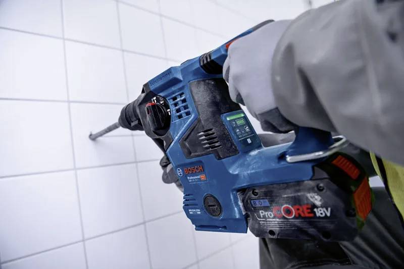 A person wearing a glove uses a Bosch ProCORE 18V power tool to drill into a tiled wall, indicating a home improvement or construction task.