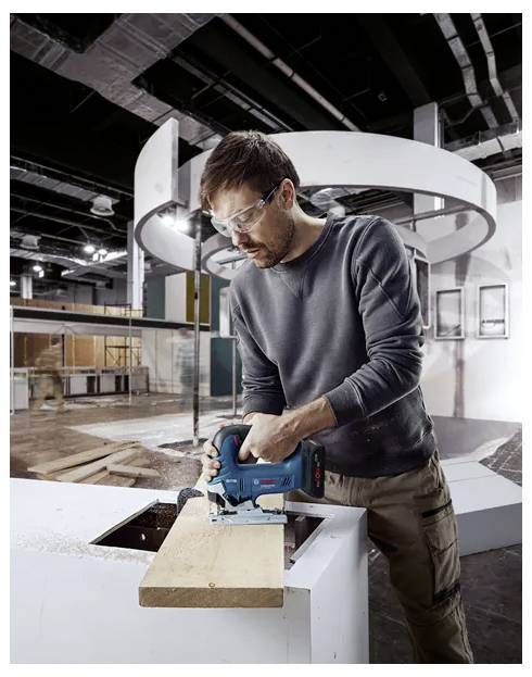 A man wearing safety glasses operates a power saw, cutting a wooden board in an industrial workshop setting.