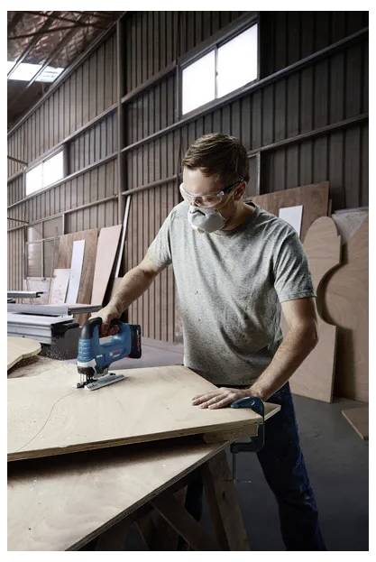 Person wearing safety gear uses a jigsaw on plywood in a woodworking shop.