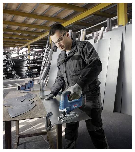 Worker in protective gear uses a jigsaw to cut a metal sheet in a factory with stacks of metal in the background.