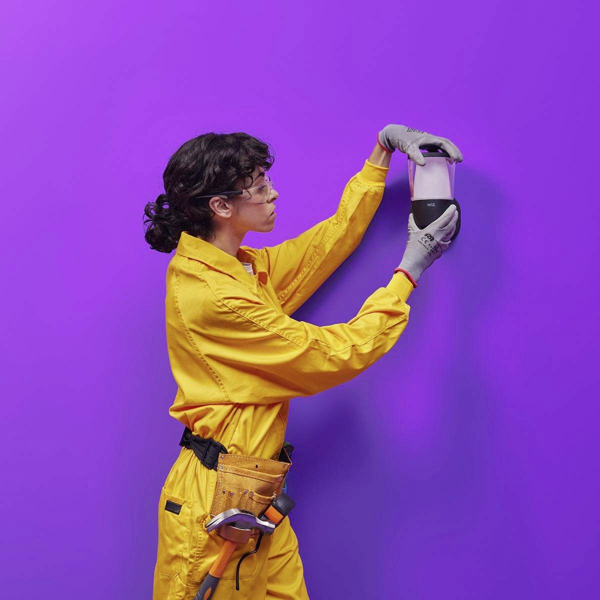 A person in a yellow overall and gloves is holding a white and black device in front of a purple background.