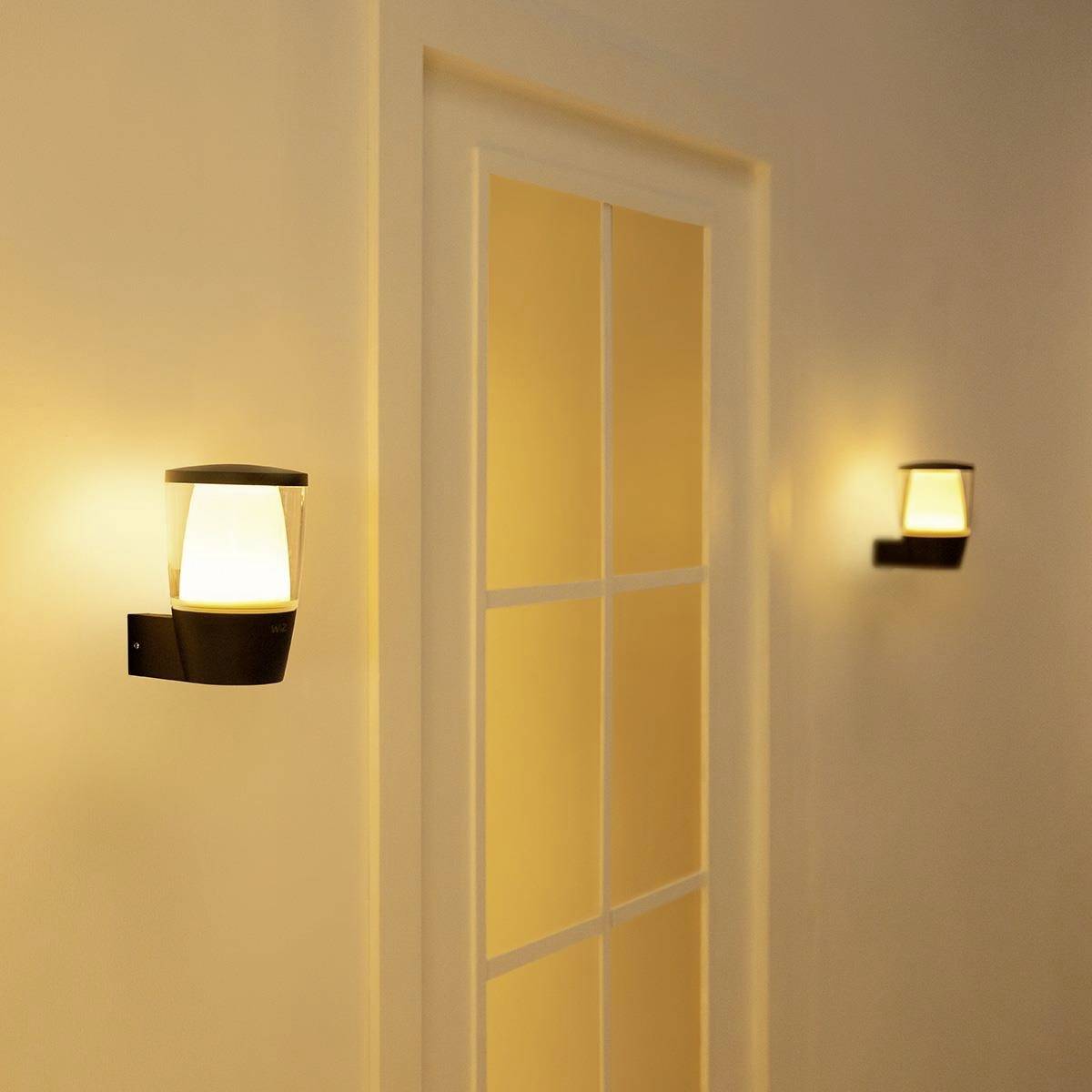 Two contemporary wall lights illuminate a hallway with a closed door frame in the centre. The lighting creates a warm atmosphere.