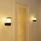 Two contemporary wall lights illuminate a hallway with a closed door frame in the centre. The lighting creates a warm atmosphere.
