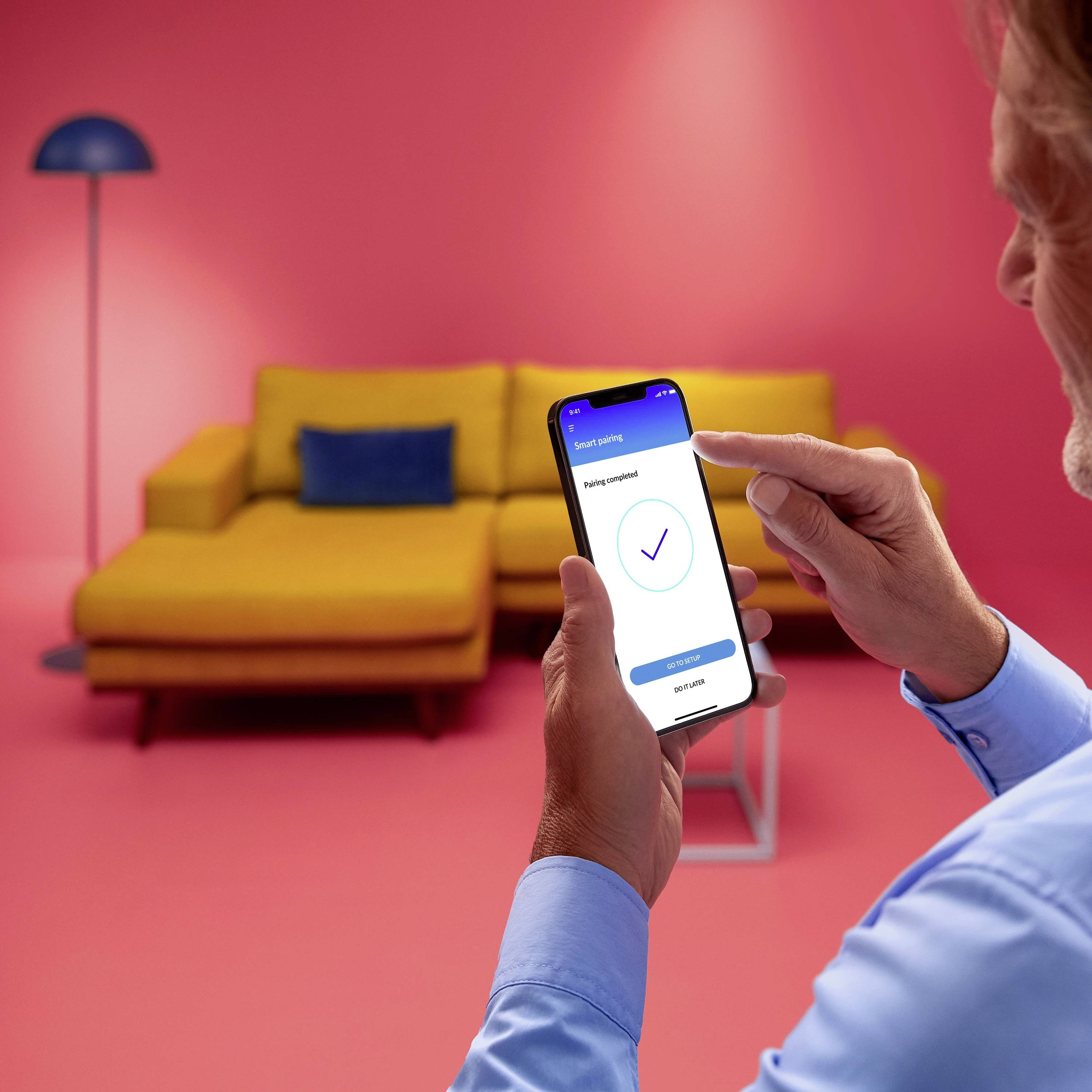 A person is holding a smartphone displaying a completed task with a tick mark. In the background, there is a yellow sofa and a blue lamp.