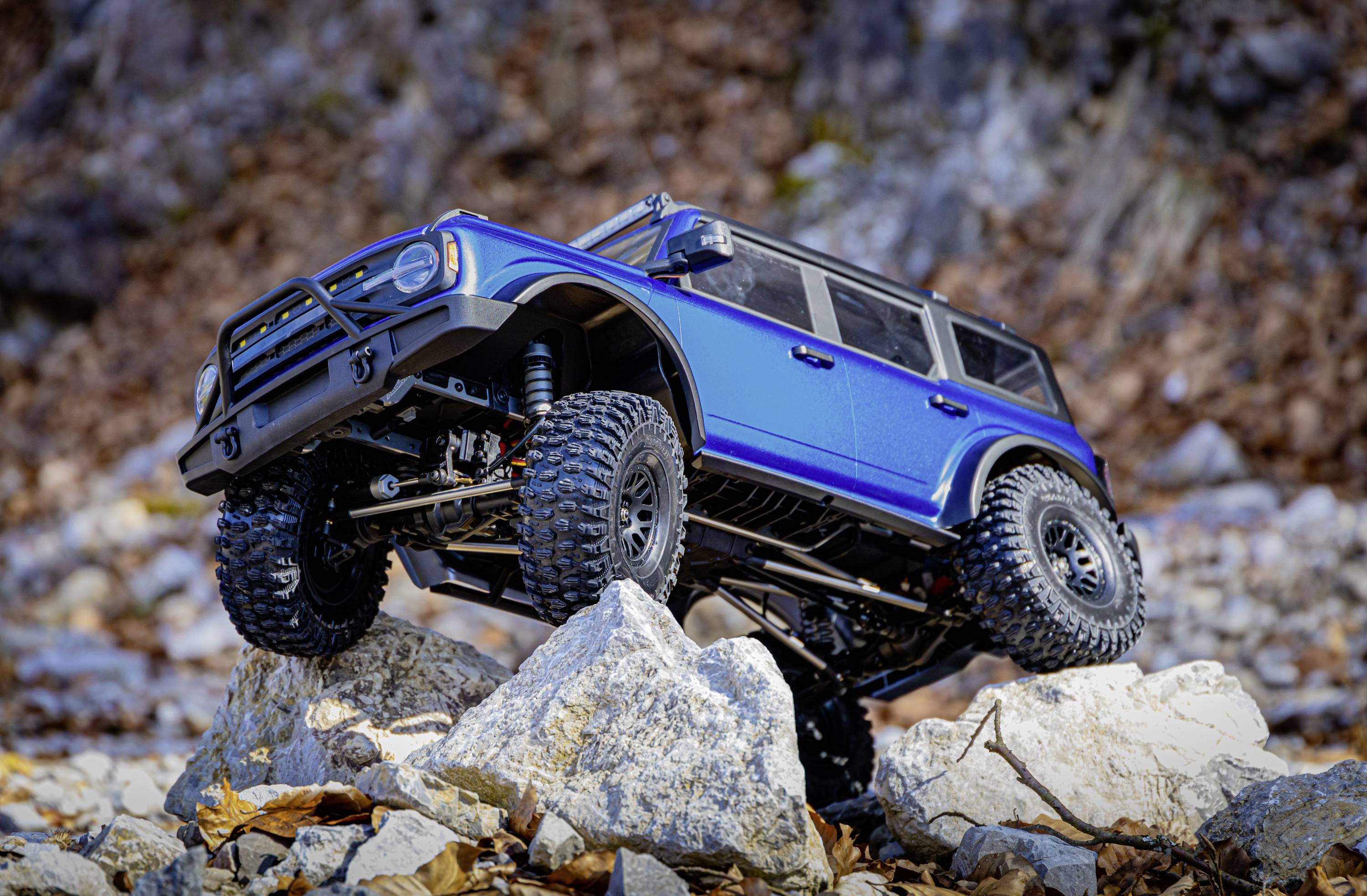 A blue off-road vehicle drives over large rocks outdoors. The background is rocky with autumn foliage. The vehicle demonstrates its off-road capabilities.