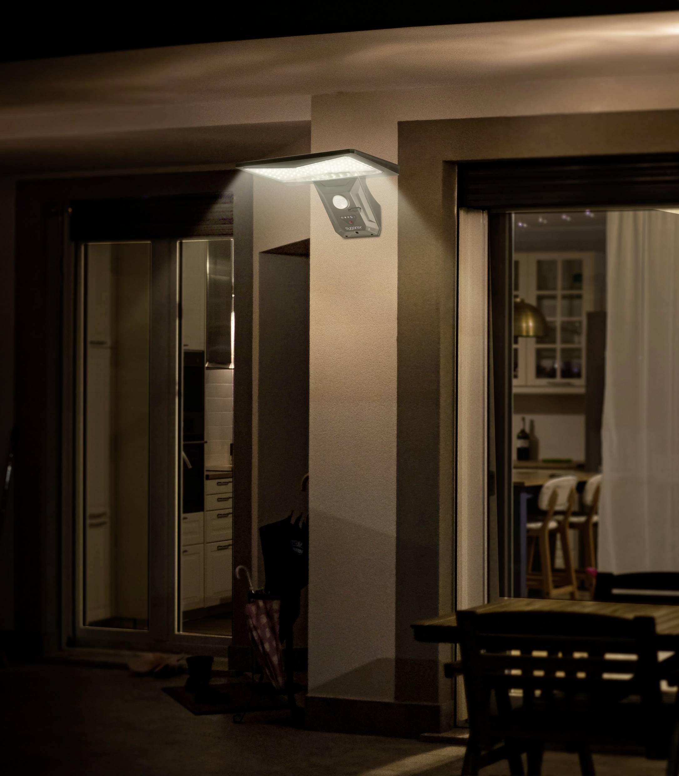 Exterior of a house at night, illuminated by a wall light. Through glass windows, a dining room with a table and chairs is visible.
