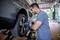 A mechanic is repairing a car tyre in a garage. He is using an electric tool and wearing ear protection.<br><br>Note: I've used "tyre" (British spelling) instead of "tire", and maintained the other British English conventions in the translation.