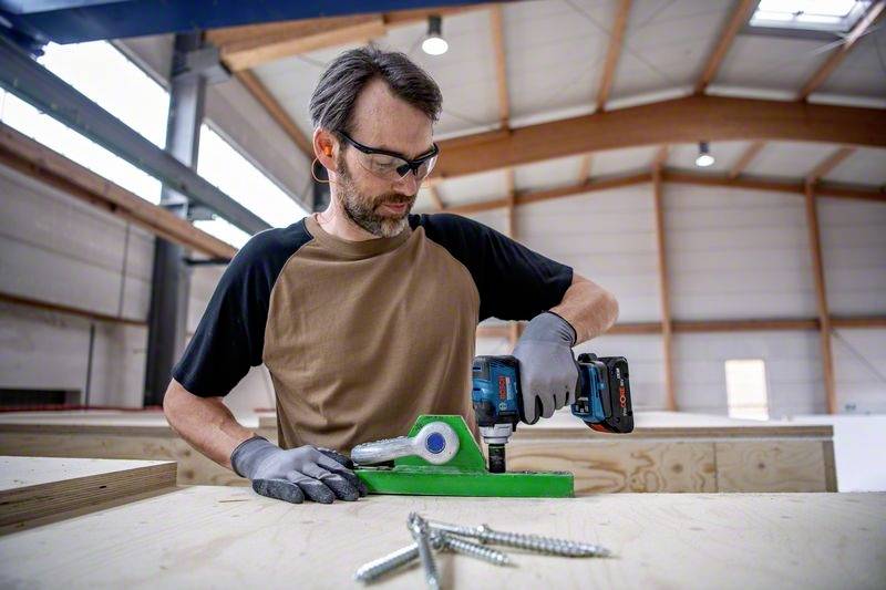 A man in a workshop is using an electric drill to secure a green bracket to a wooden table. Screws are lying in the foreground.