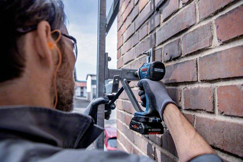 A person is installing a mounting bracket on a brick wall using a cordless drill. The person is wearing safety equipment.