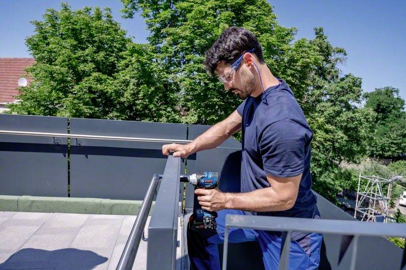 A man wearing safety glasses is working on a balcony and using an electric drill to secure something. Background: green trees.