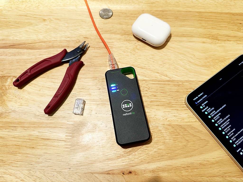 On a wooden table lies a network tester with an orange cable plugged in, alongside AirPods, a pair of pliers, and a tablet.