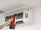 A hand is holding a red and black device in front of an open air conditioning unit for cleaning or inspection. A ceiling is visible.