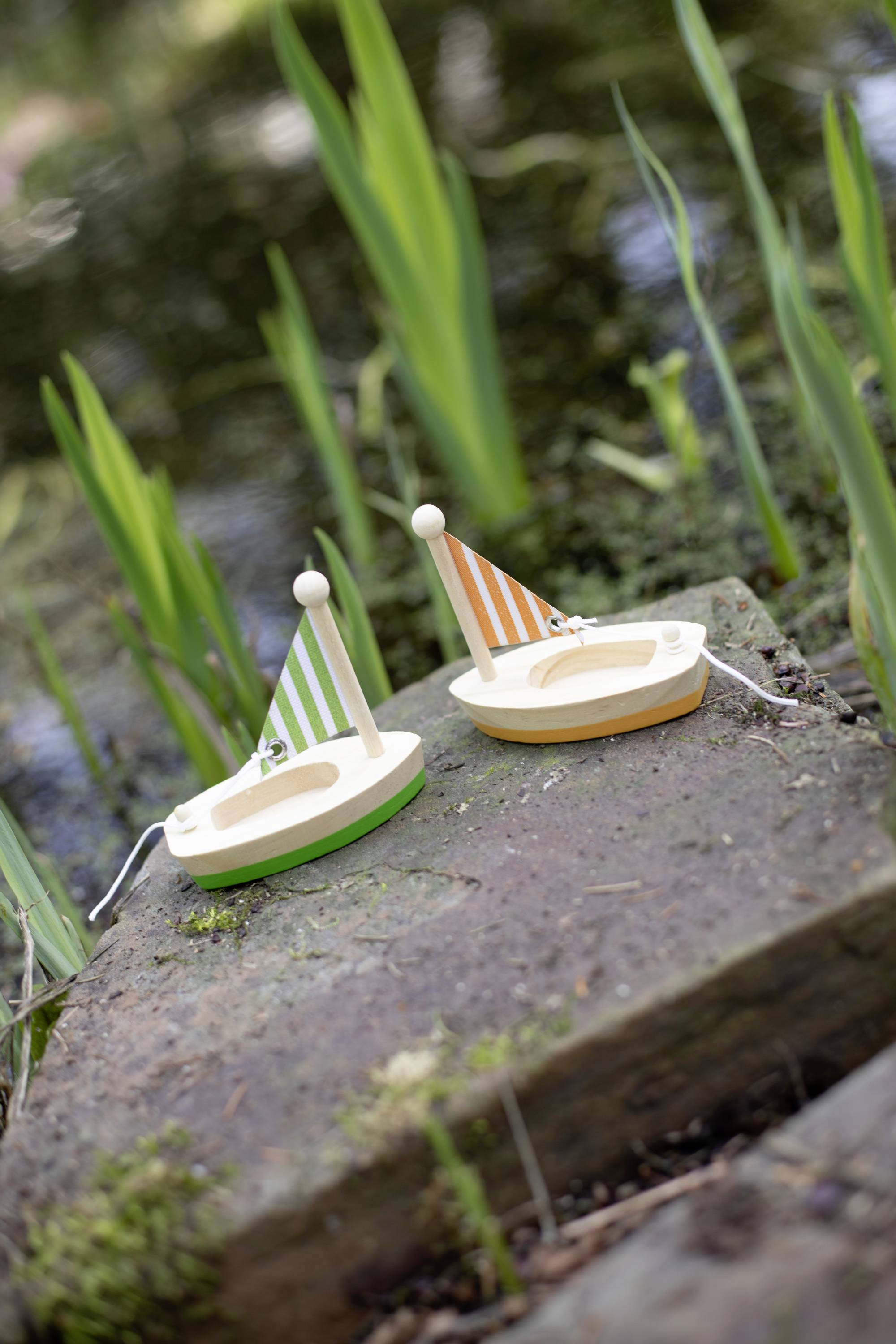 Two small wooden sailing boats with striped sails, one green and white and the other orange and white, are positioned on a rock by the pond's edge.
