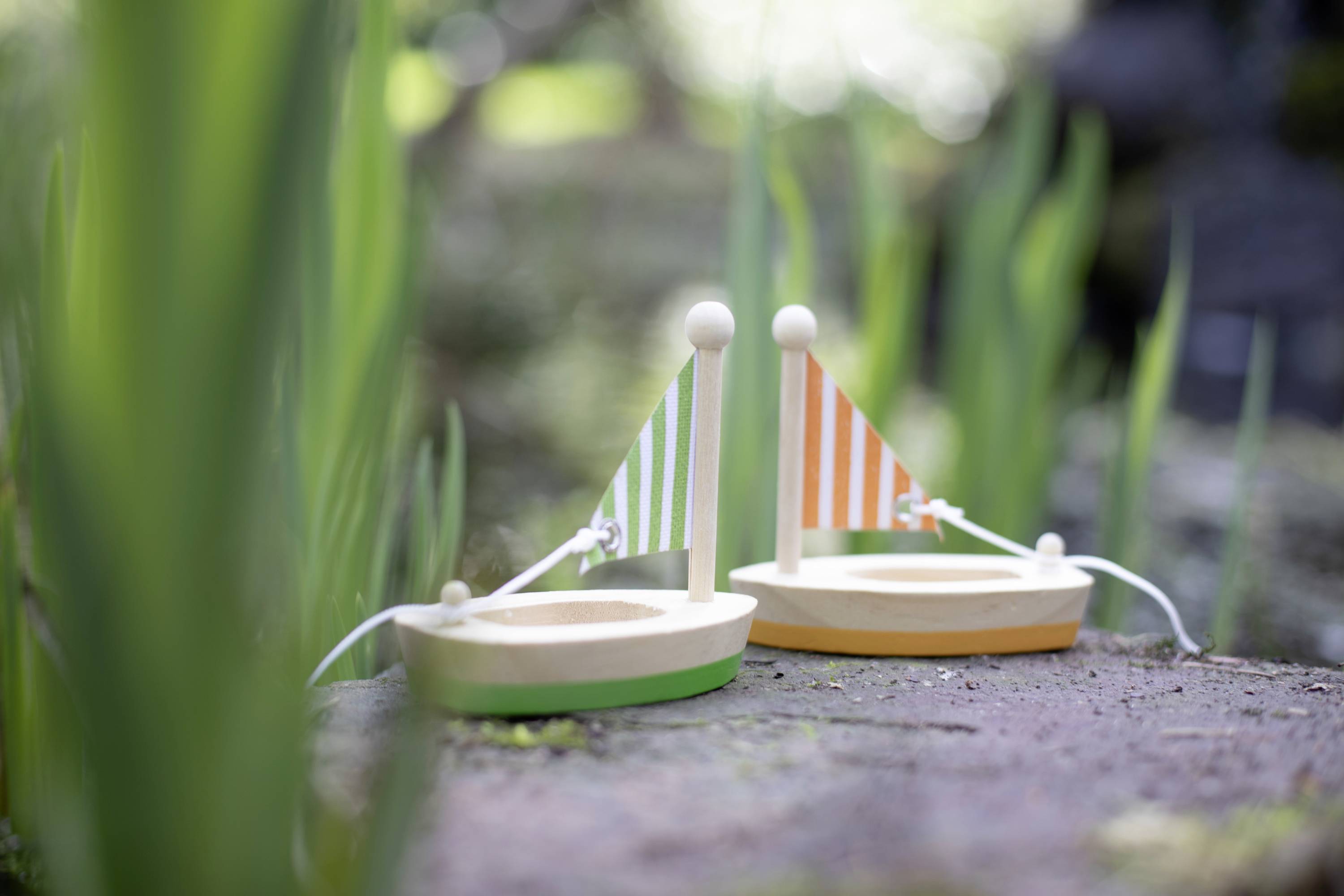 Two small wooden boats with colourful sails sit on a stone, surrounded by green plants.