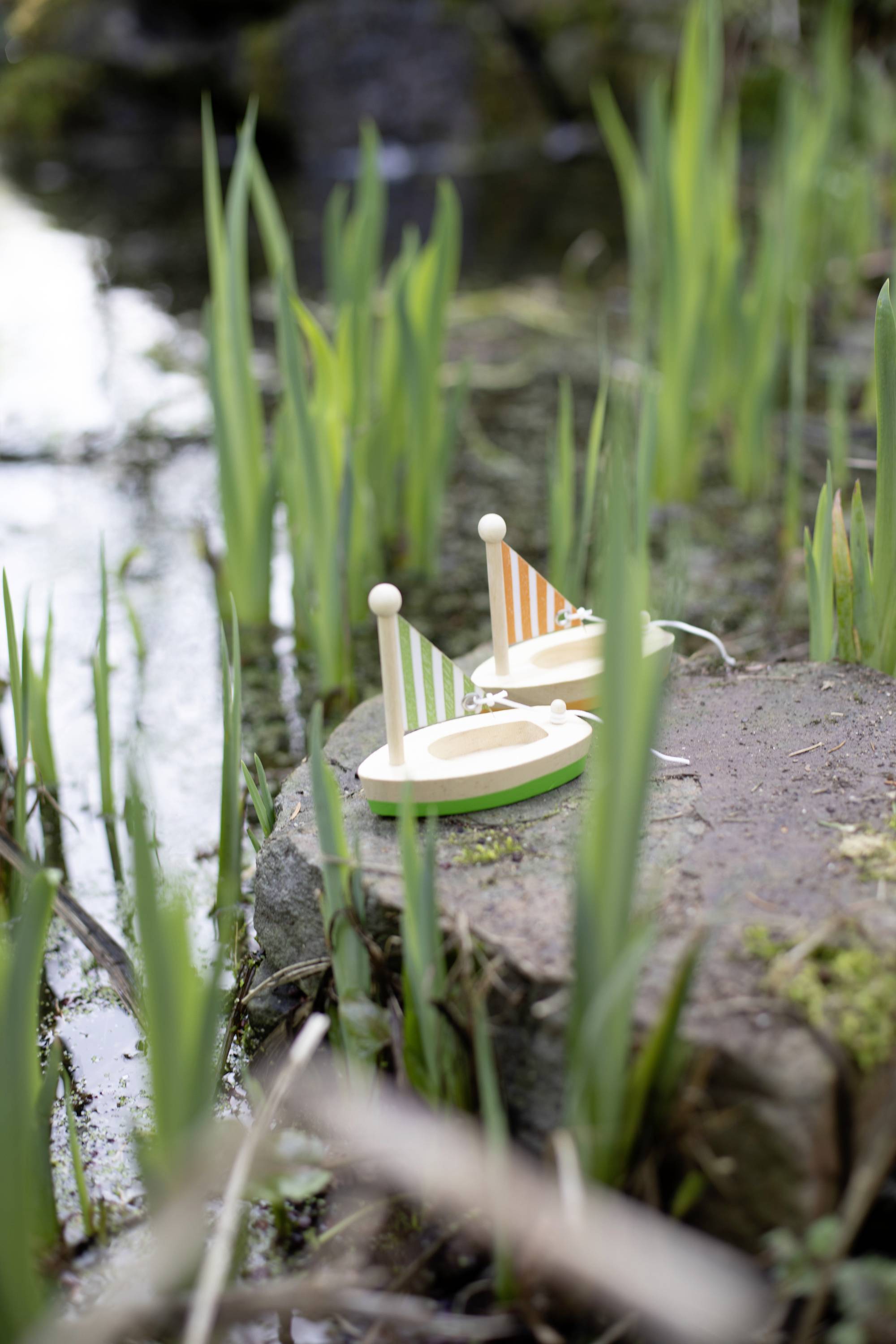 Two small wooden boats with striped sails sit on a stone at the edge of a grass-covered pond.