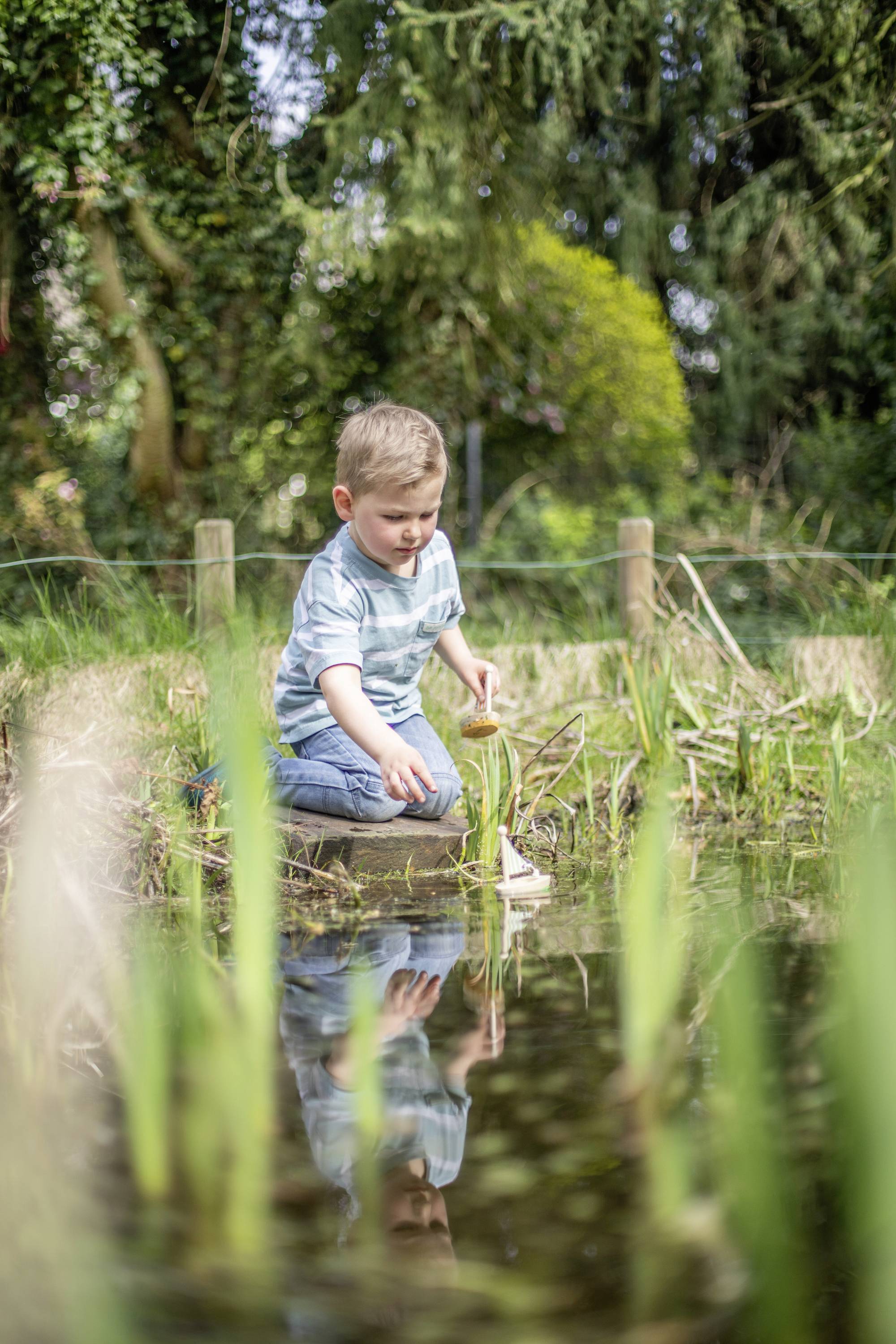 A boy in a striped shirt kneels by the pond's edge and plays with a model boat in the water. The background shows green vegetation.