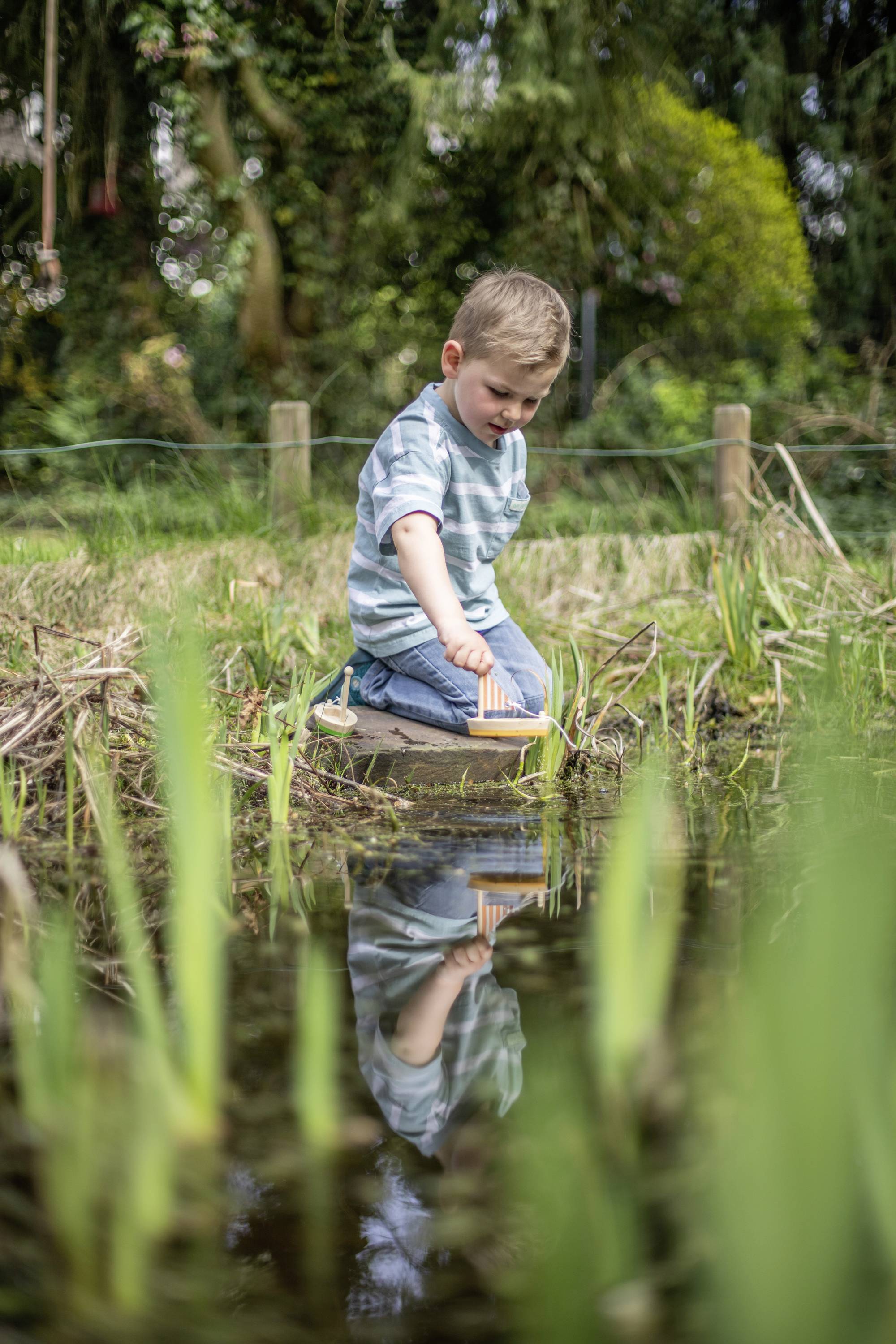 A child kneels at the pond's edge in the grass and plays with a small wooden boat in the water. Trees and plants are visible in the background.