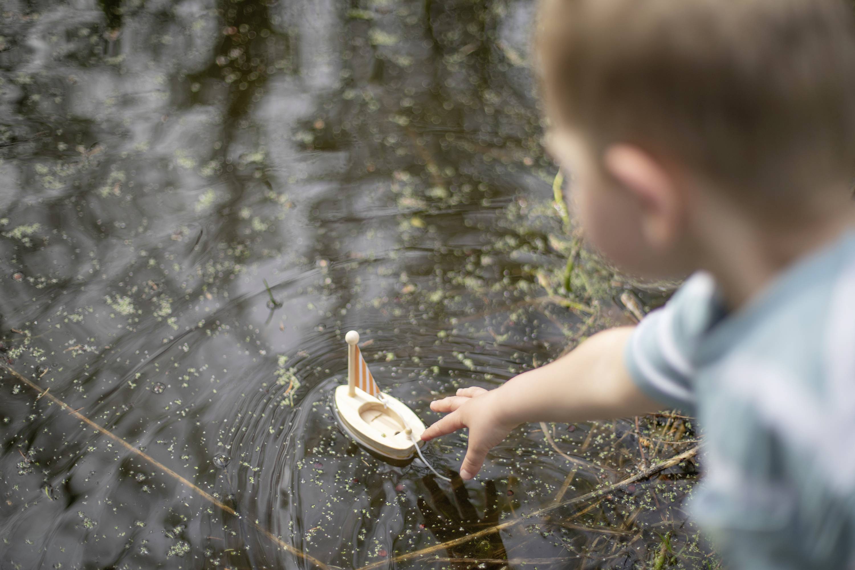 A child looks at a small wooden toy boat floating in the water. The child stretches out their hand to reach the boat.
