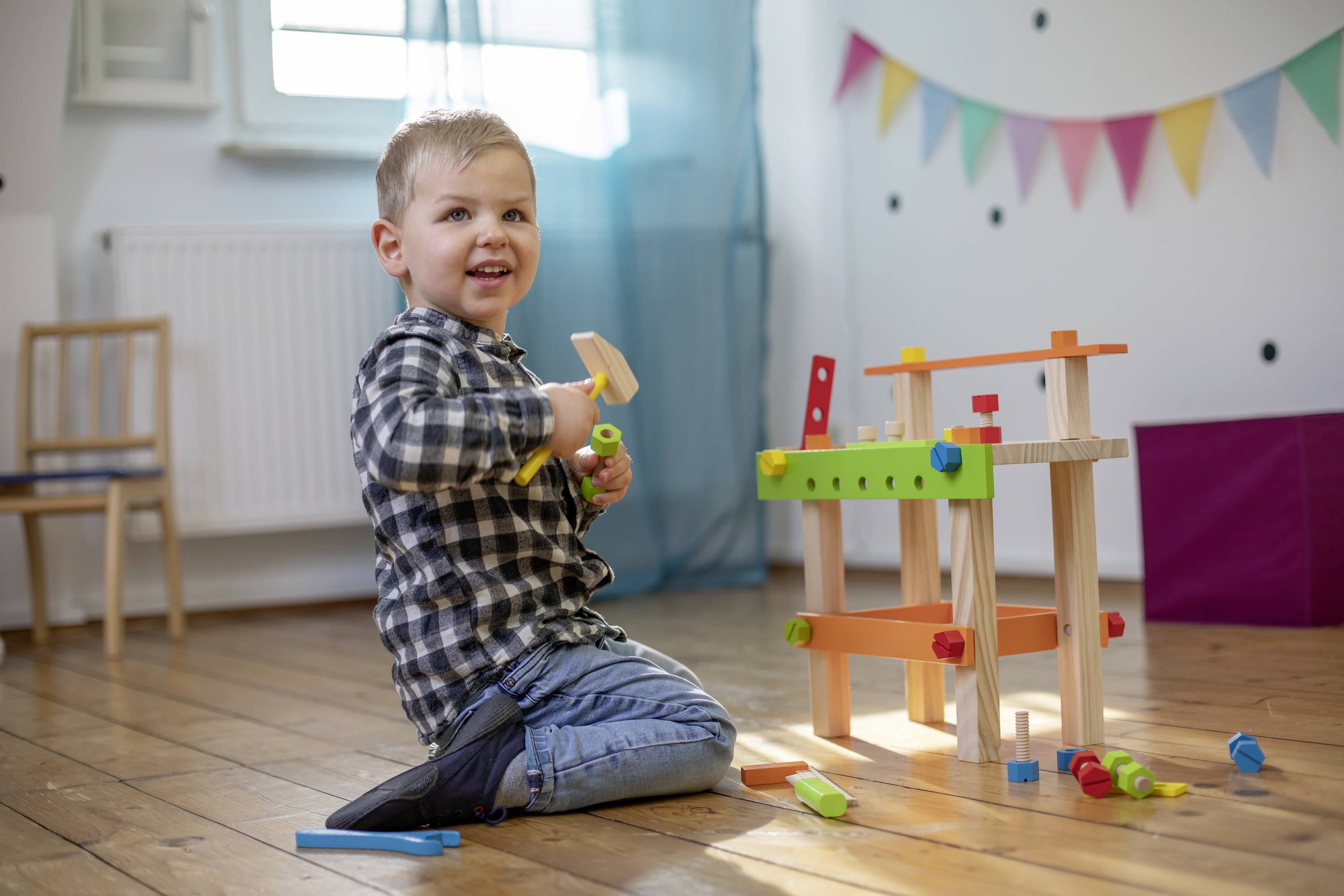 A smiling child sits on the floor playing with a colourful wooden workbench. Colourful bunting hangs on the wall in the background.
