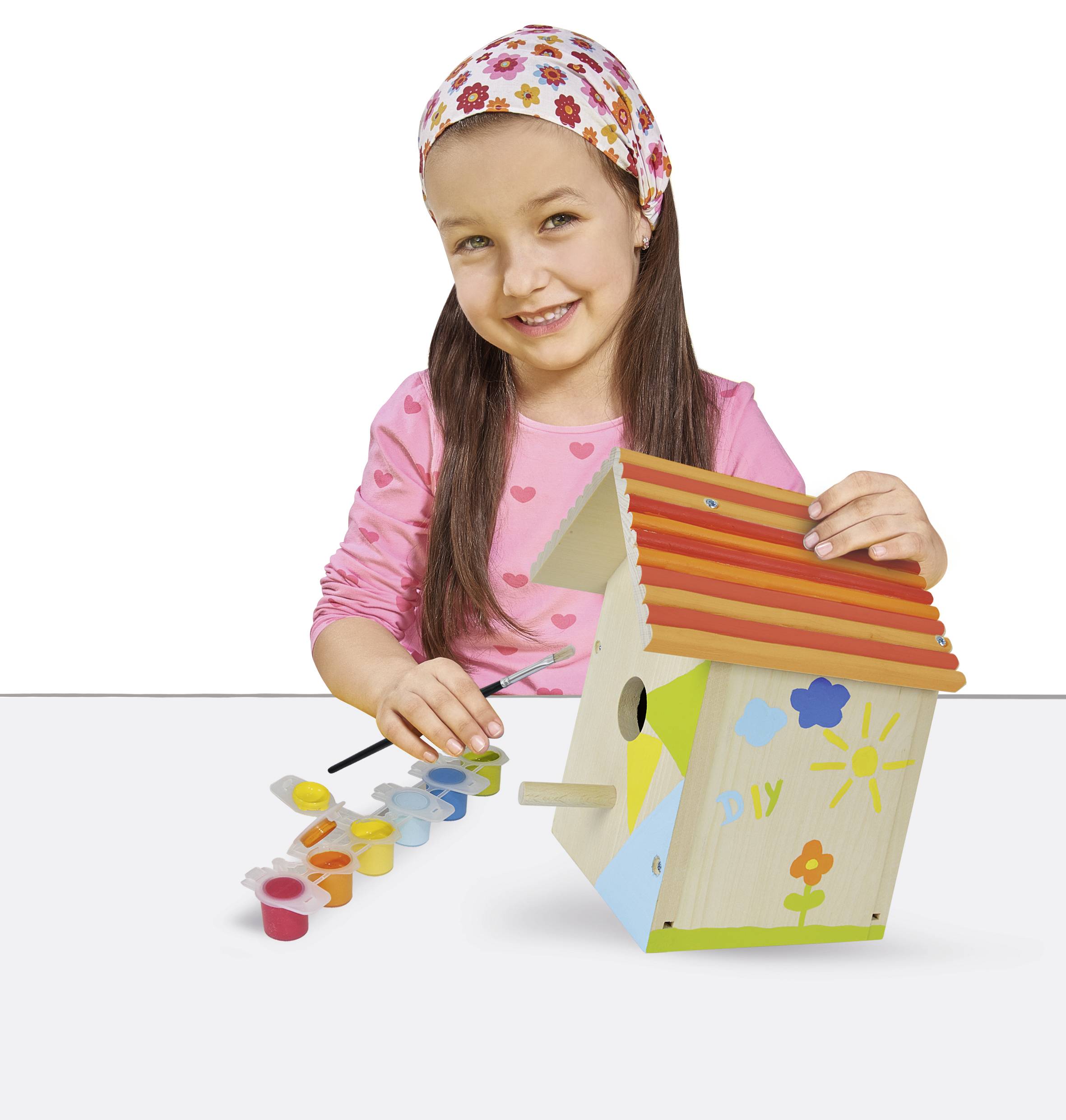 A child is painting a birdhouse in bright colours. They are wearing a pink top and a flowery headband. Various paint pots are on the table.