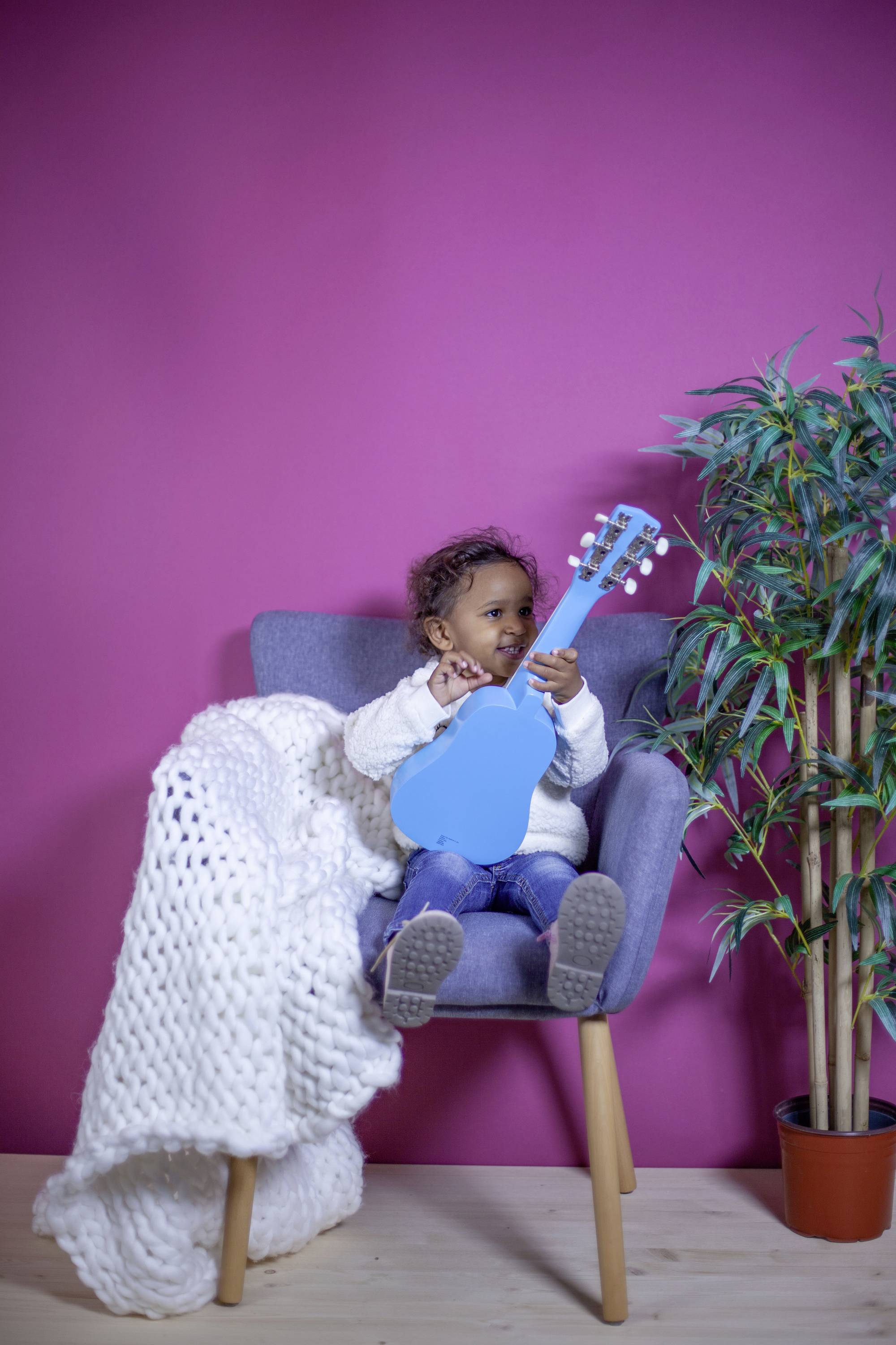 A toddler sits on a chair with a blue toy guitar against a pink background, next to a white knitted blanket and a plant.