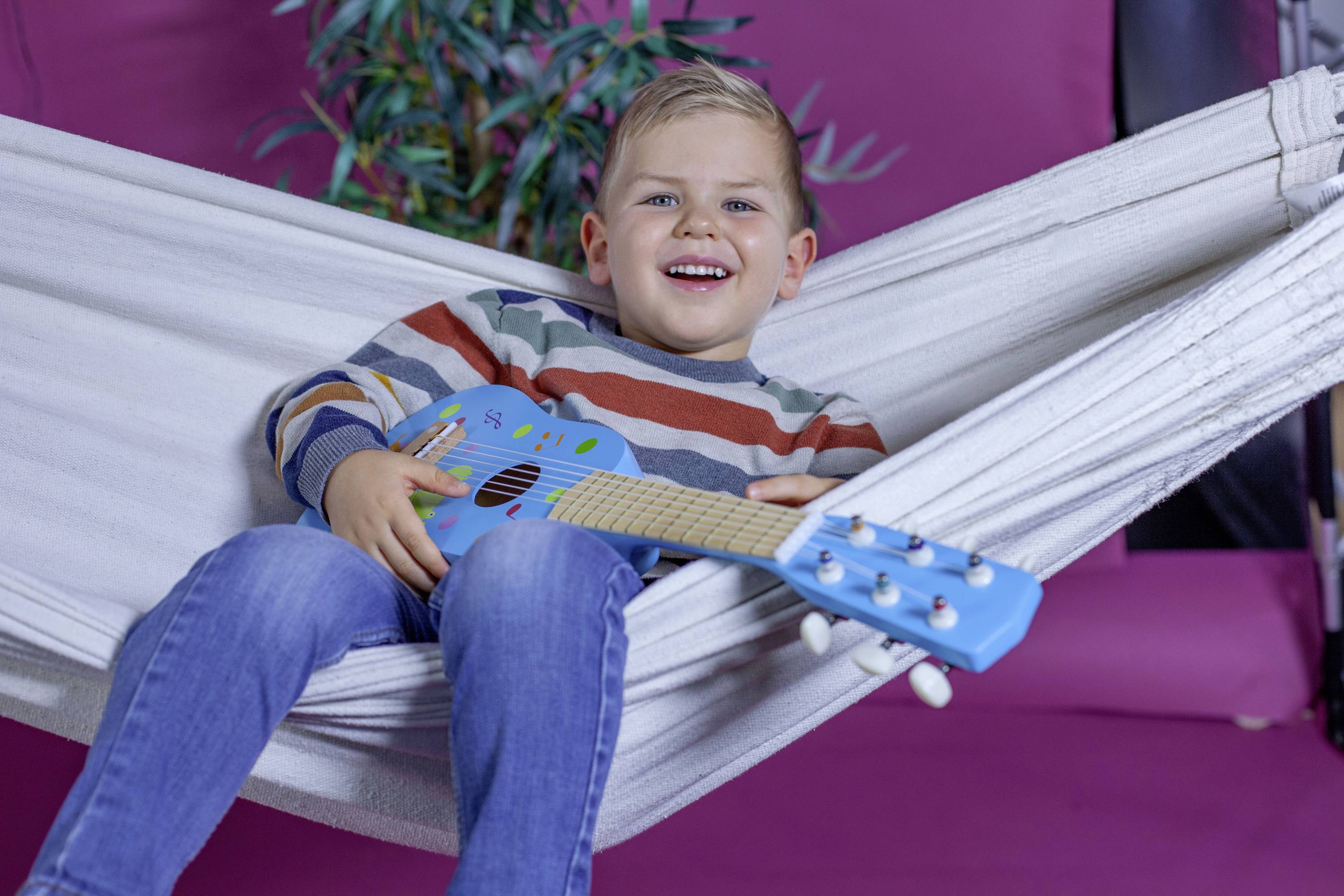 A smiling child is lying in a hammock and playing a blue guitar. In the background, there is a plant.
