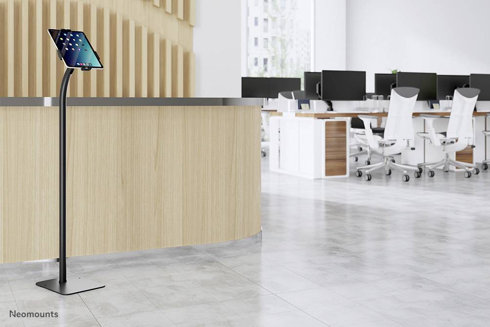 Tablet on a tall stand next to a modern reception desk in an office with white desks and chairs in the background.