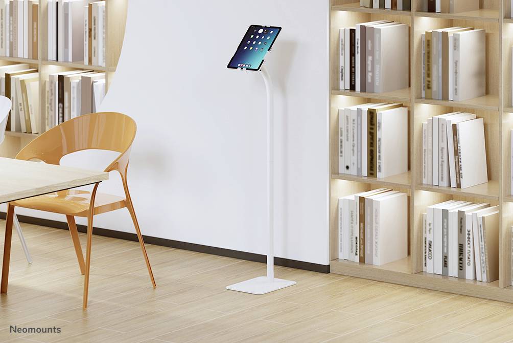 A tablet is resting on a white stand next to an empty orange chair in front of shelves with books in a modern room.