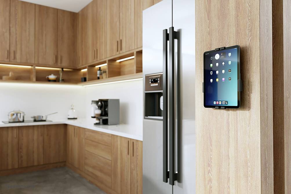 A tablet mounted on a wooden wall in a modern kitchen, displaying app icons. A fridge and kitchen worktops are visible in the background.