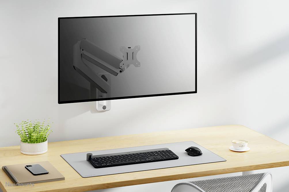 Flat monitor arm mounted on the wall, above a minimalist desk with keyboard, mouse, notebook, plant and cup.