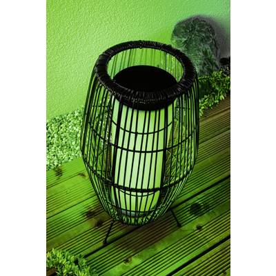A black, round lantern with green light stands on a wooden floor. In the background are green pebbles and an illuminated wall.