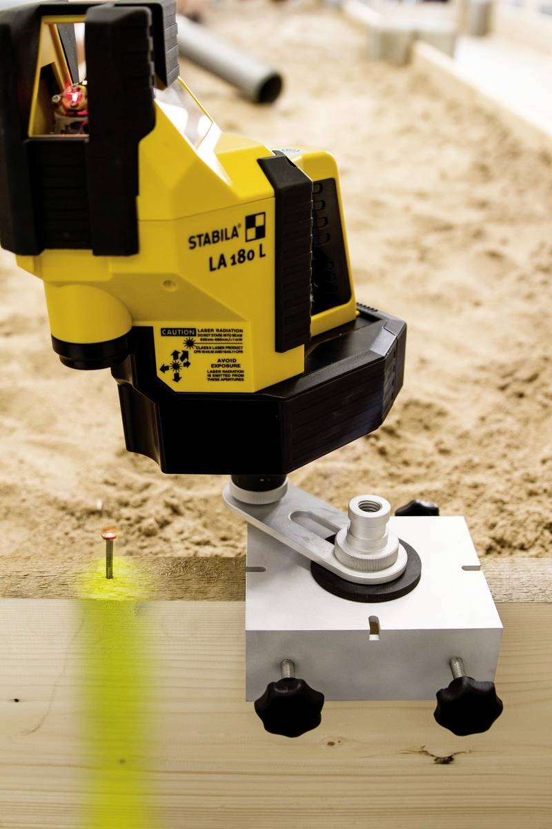 A yellow, laser-based precision measuring device aligns a laser beam towards a wooden frame, with sand in the background.