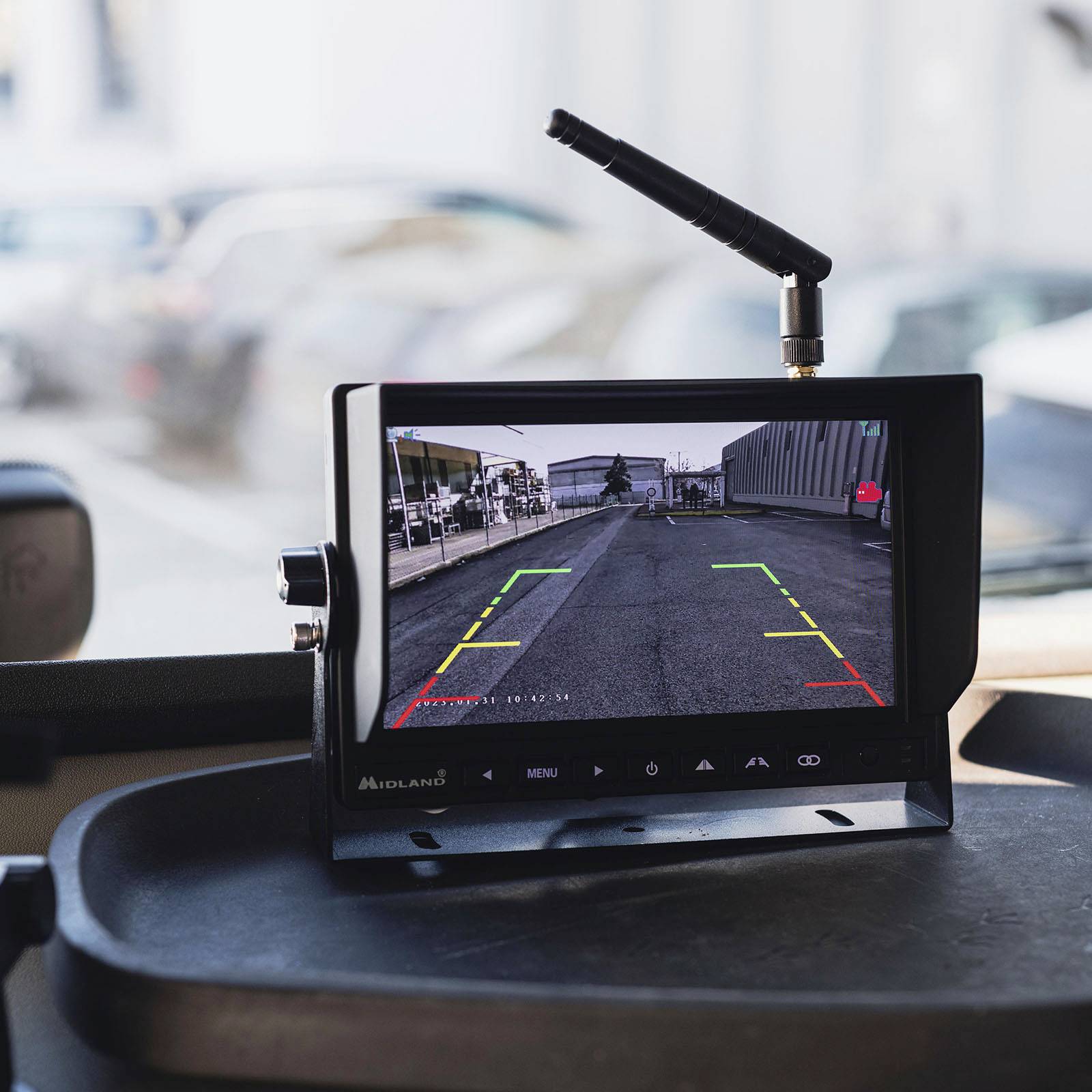 Screen in a vehicle displays the reversing camera with coloured lines for navigation. Blurred buildings are visible in the background.