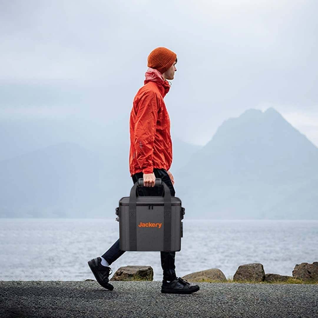 A person wearing a red jacket and hat carries a black 'Jackery' bag by the sea with misty mountains in the background.