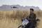 A man sits on a motorbike in a vast grassland, controlling a drone. Rocky formations are visible in the background.
