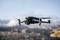 A black drone flies in the air above a blurred urban landscape. It has four rotors and a camera on its front.