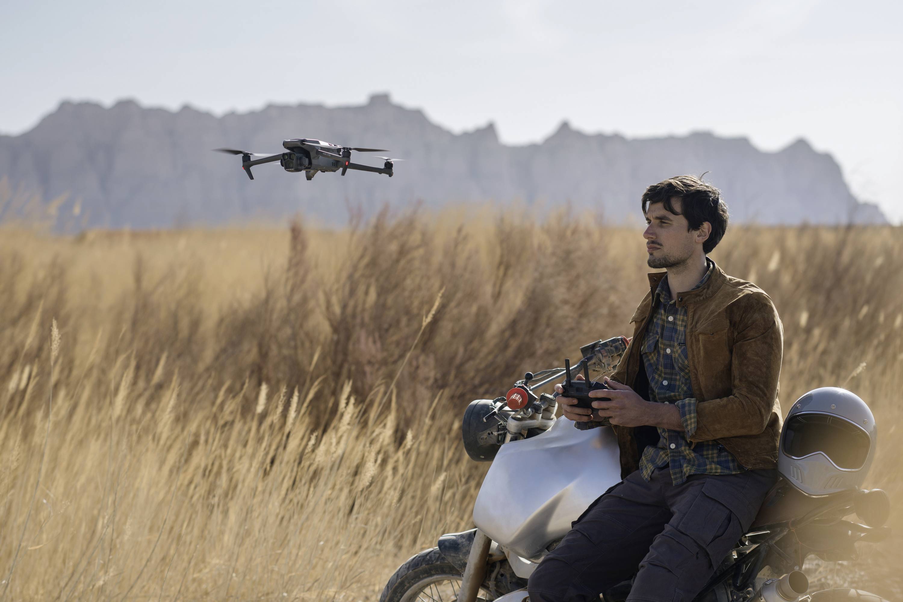 A man sits on a motorbike, controlling a drone over golden fields with a mountainous backdrop. Relaxed and focused.