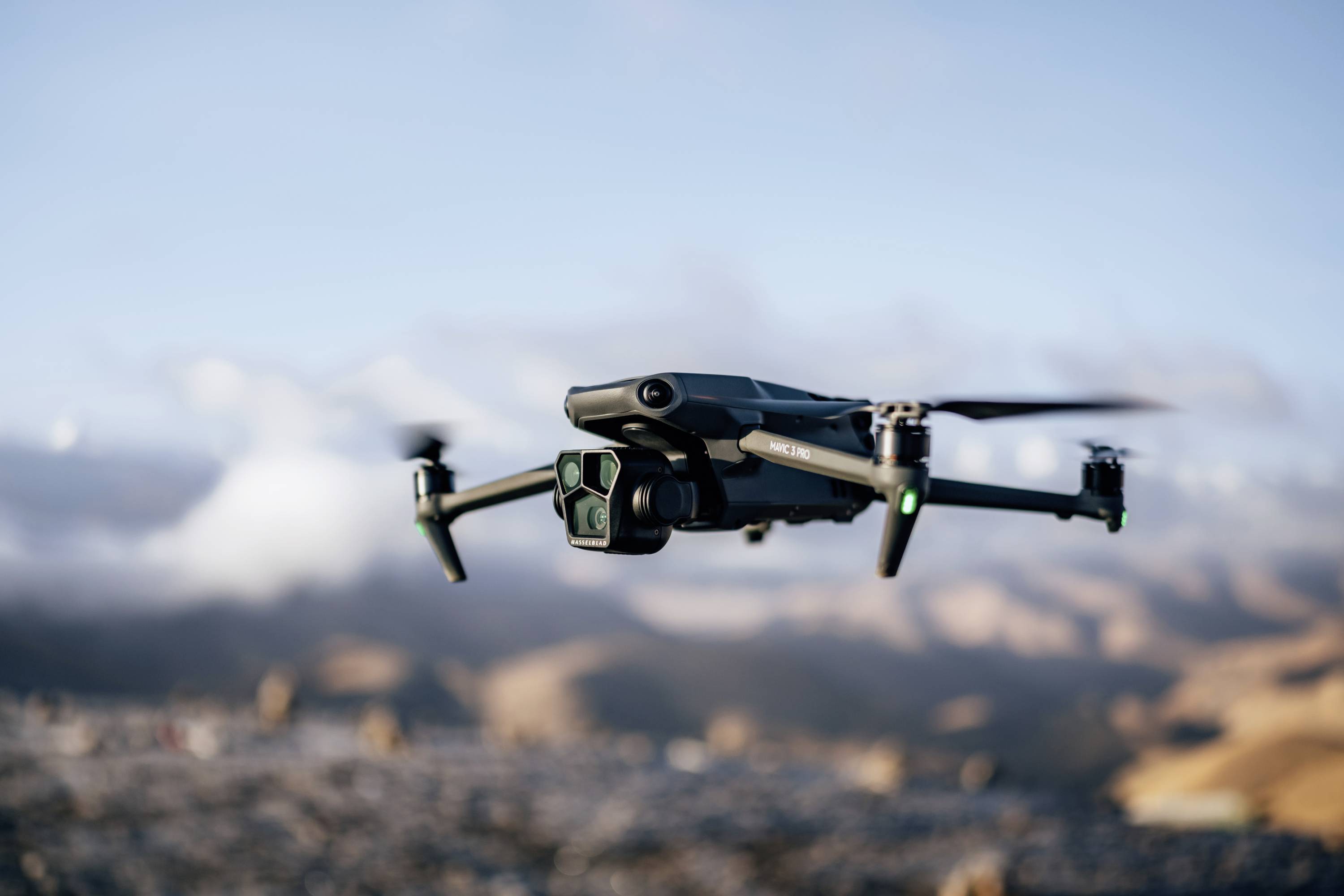 A drone flies over a landscape with mountains and clouds in the background, under a clear sky. It is equipped with a camera.