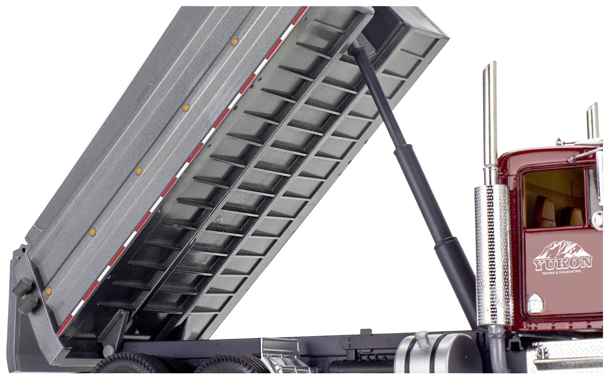 Tipper truck viewed from the side. The load bed is raised, showing its stability structure. Red-painted driver's cab with Yukon logo.