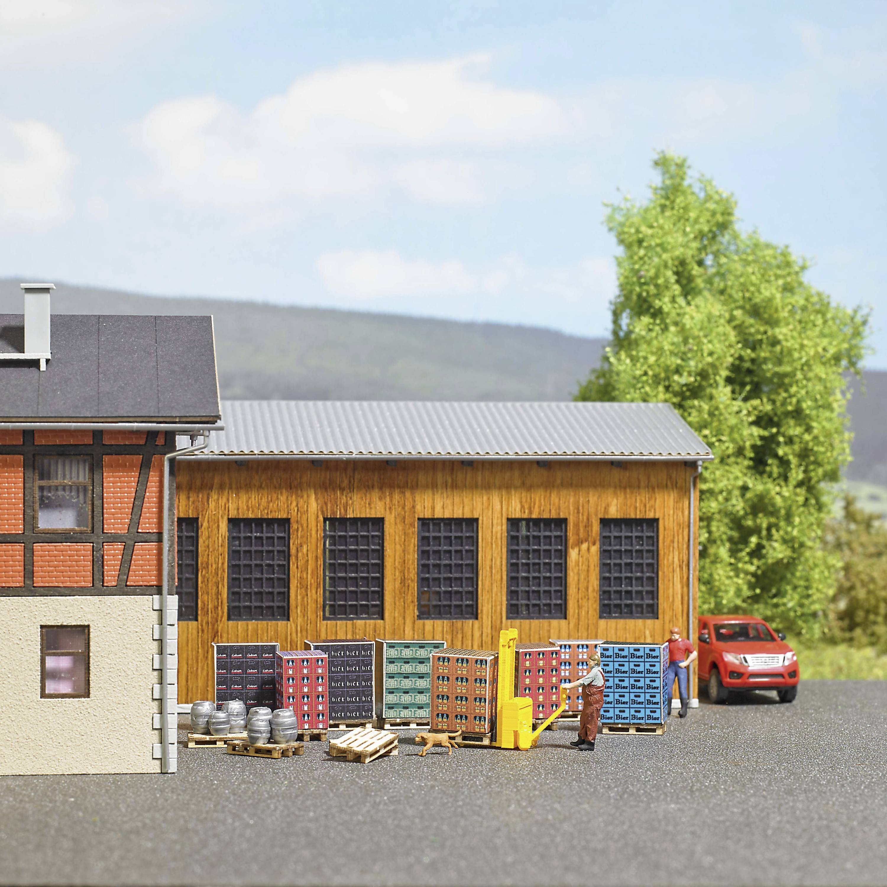 A brick warehouse with red bricks and a grey roof. In front of the building are stacked pallets, barrels, and a red delivery van.
