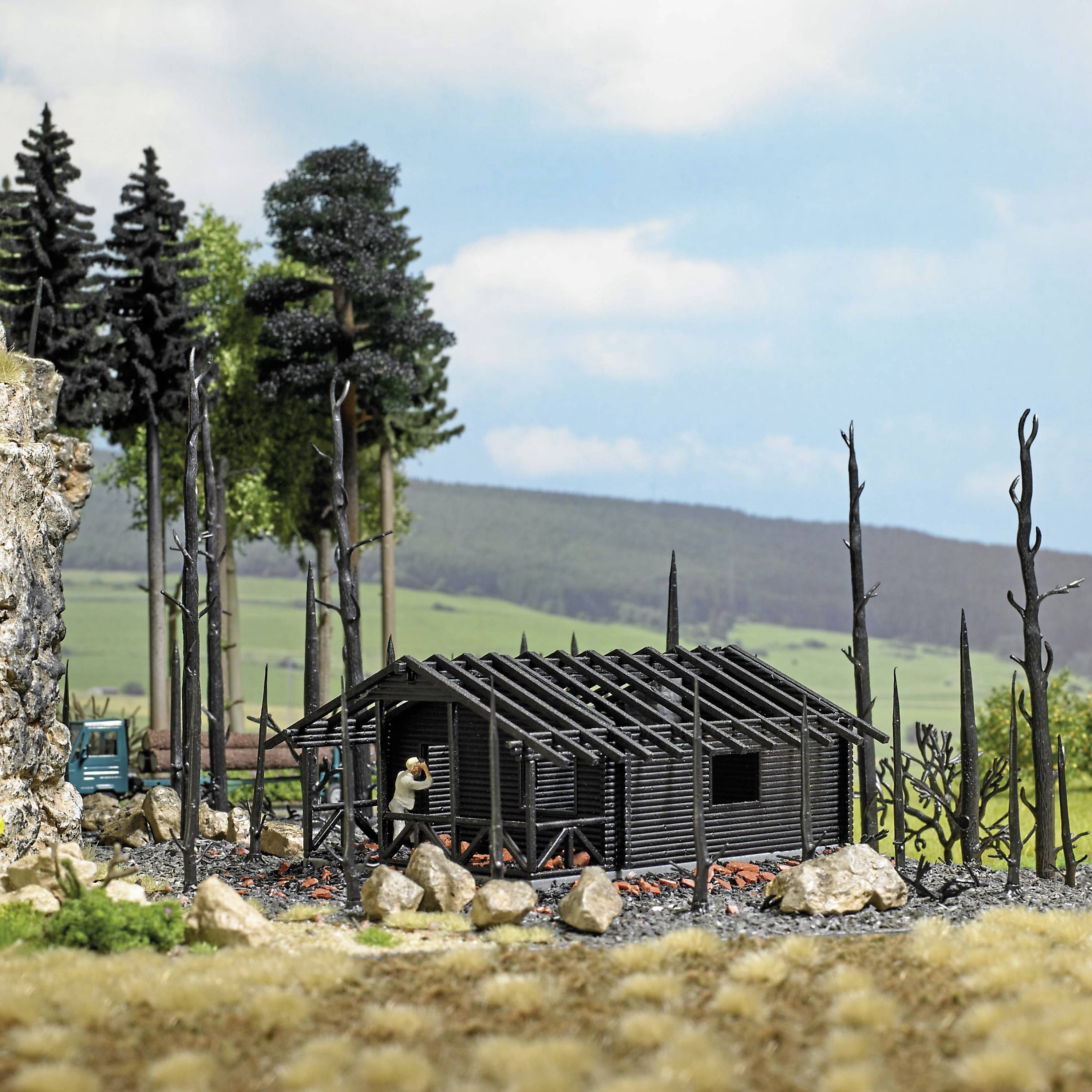 A small model house under construction in a rural setting, surrounded by trees and rocks, beneath a cloudy sky.