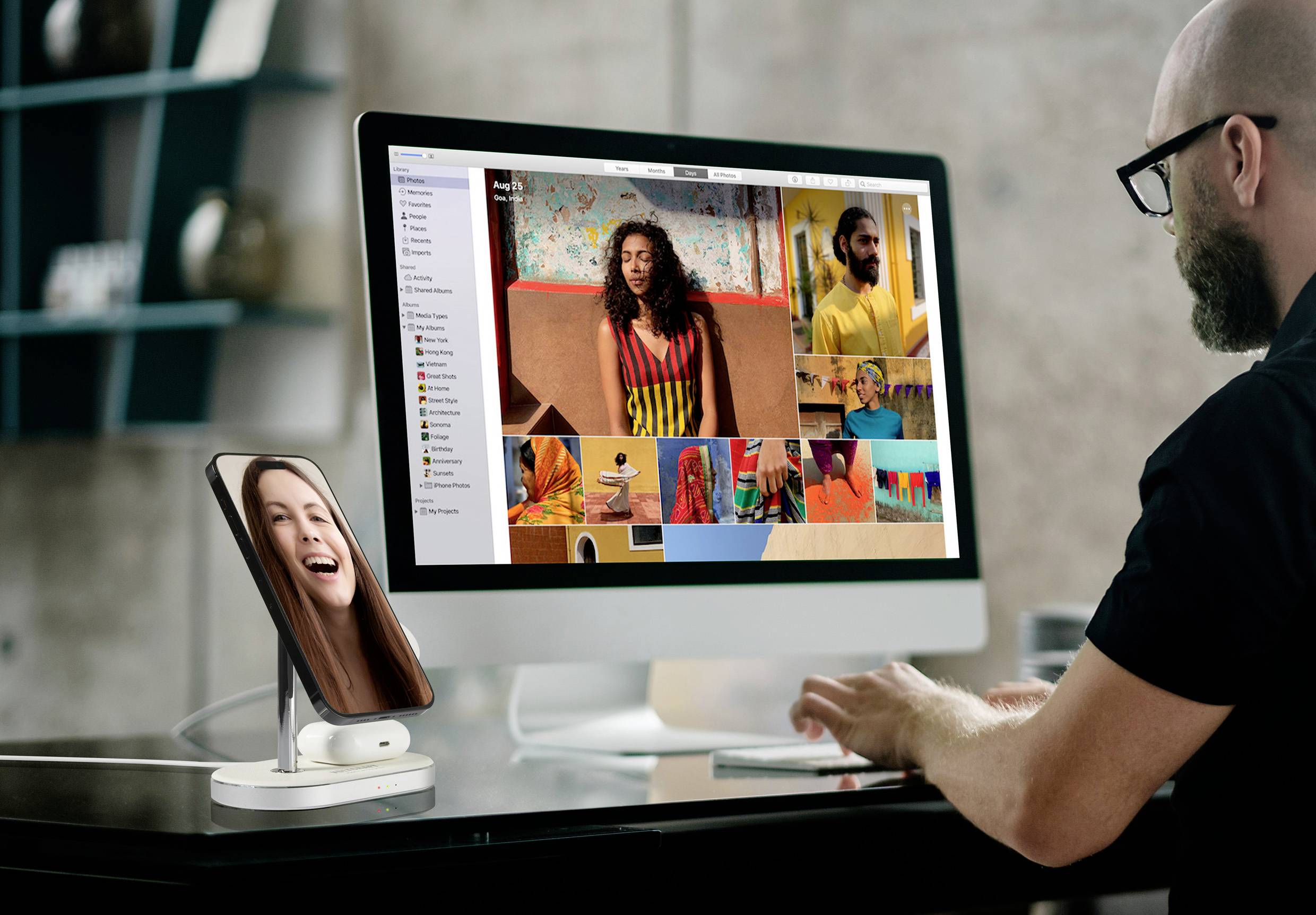 A person is working on a computer with several photos displayed on the screen. A smartphone shows a smiling person on a video call.