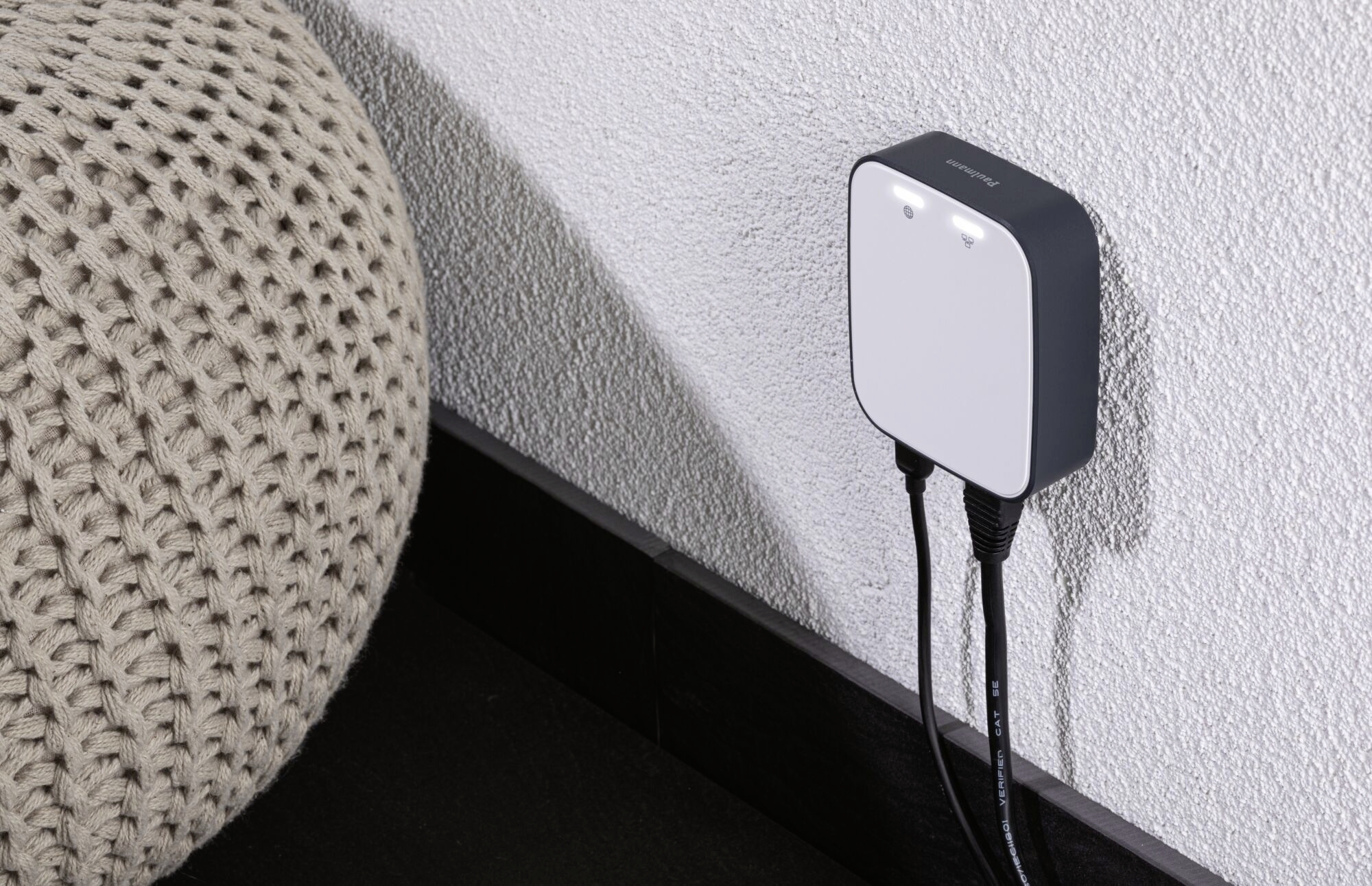 A white charger with two connected cables is plugged into a wall socket. To the left of the image is a knitted pouf.