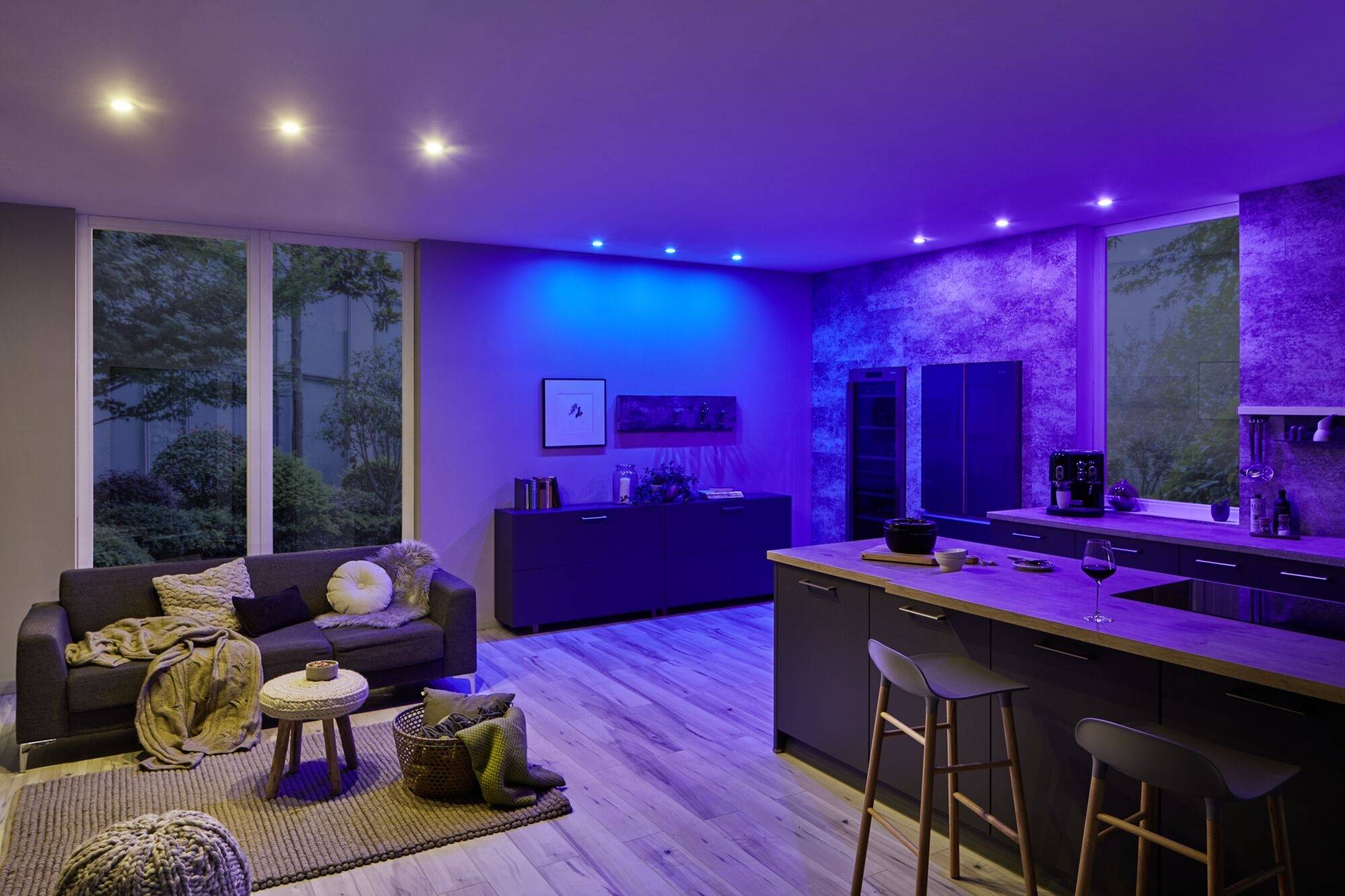 Modern kitchen and living room with blue LED lighting, grey sofa, wooden flooring and large windows. Cosy, stylish atmosphere.