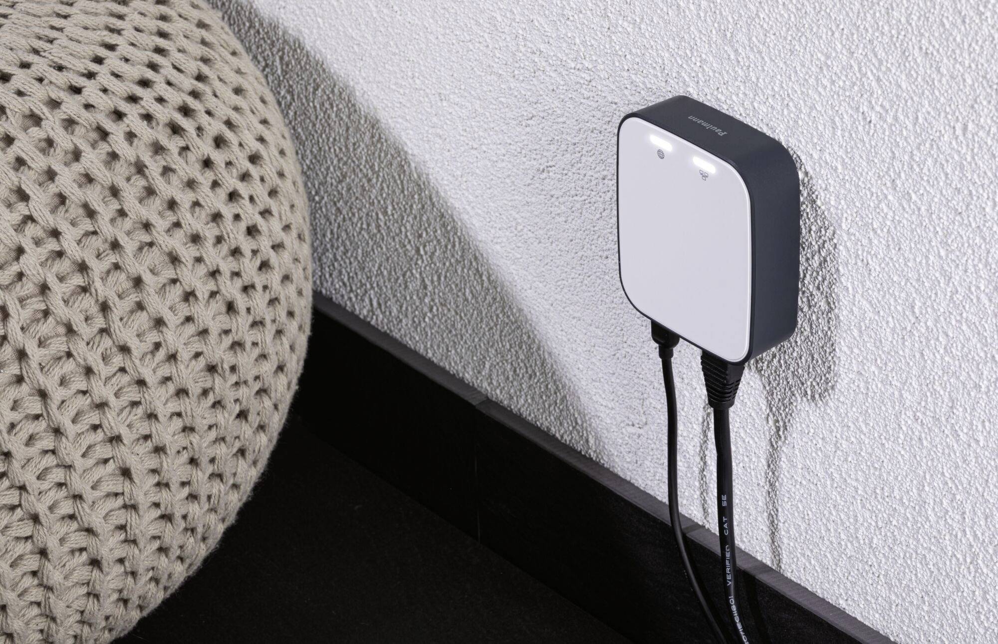 A smart device is plugged into a wall socket. Two black cables lead away from it, and it is located in a modern living space.