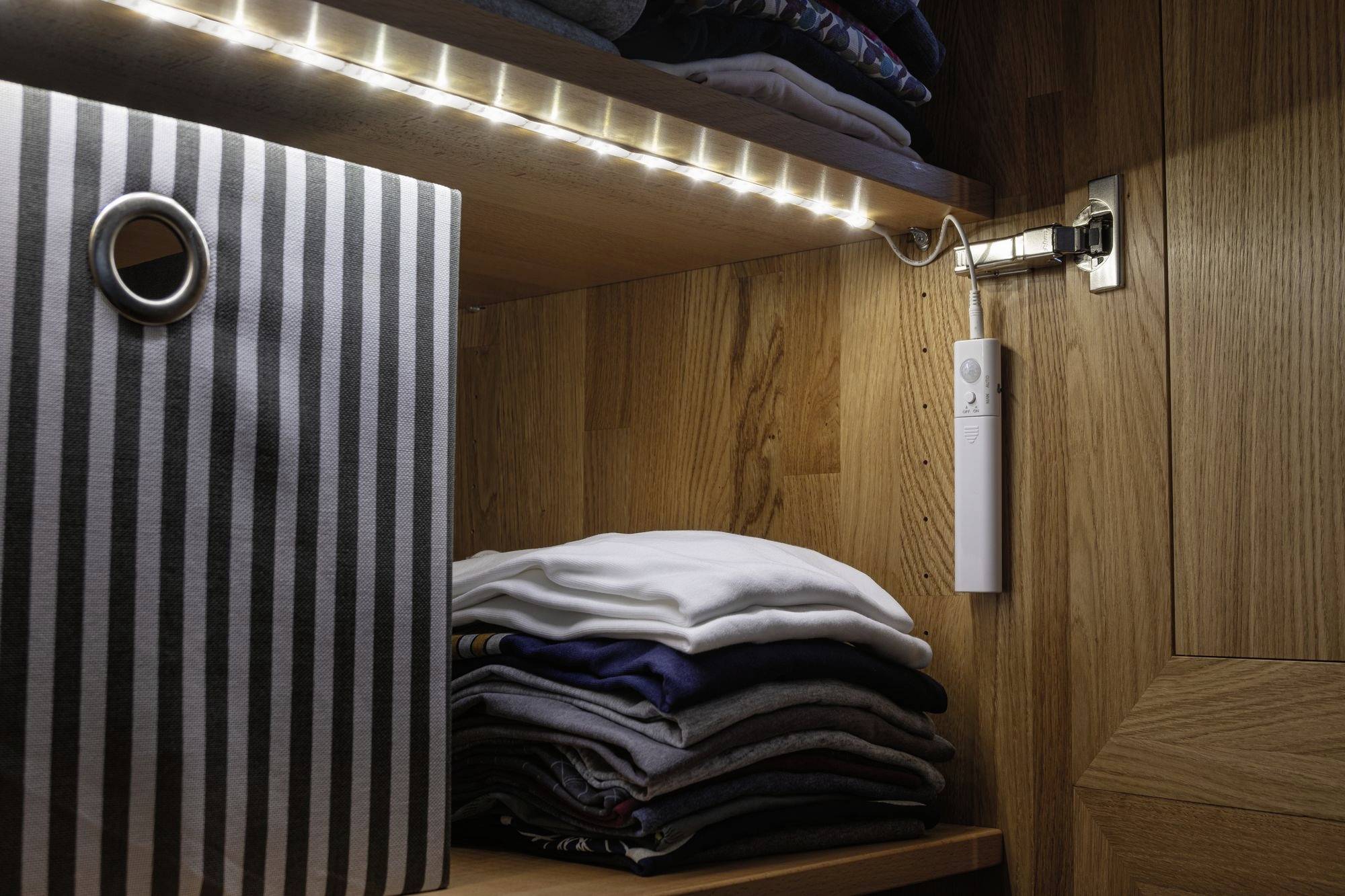 A wooden wardrobe with folded clothing on a shelf and a striped container. An LED strip illuminates the interior.