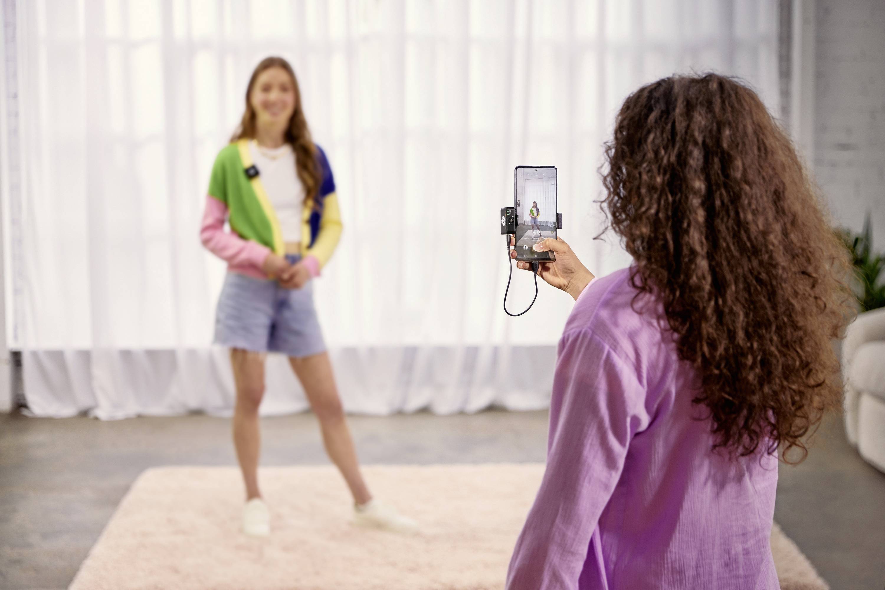 A person is photographing another person who is posing in casual clothing in a bright room in front of a curtain.